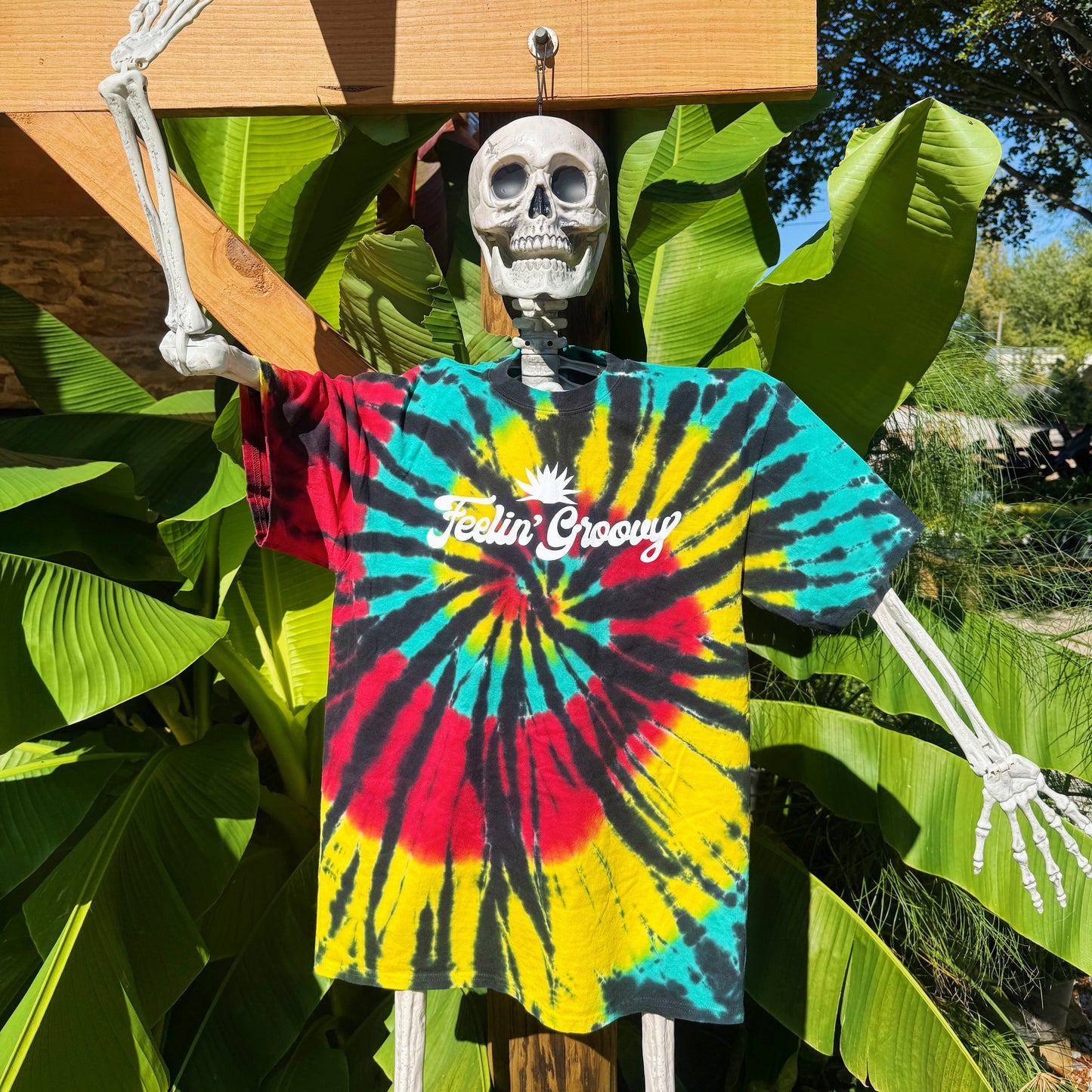 Skeleton wearing a tie-dye shirt with 'Feelin' Groovy' text, surrounded by green plants.